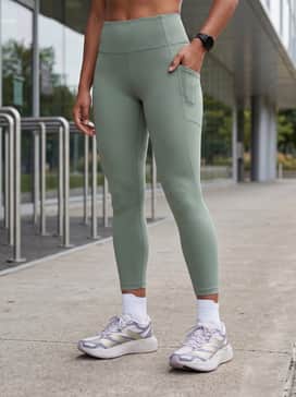 An elegant tonal gym look consisting of sage green high-compression leggings, a matching sports bra, and a sheer white technical mesh overlay top.