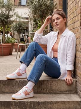 Straight-leg blue jeans with a white button-down shirt left open over a peach camisole, finished with gold minimalist jewelry to match the rose gold swoosh.