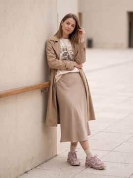 A neutral-toned beige midi slip skirt contrasted with a tucked-in oversized graphic t-shirt and a lightweight tan trench coat for a sophisticated yet sporty streetwear vibe.