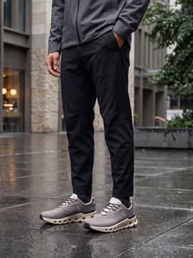 A rainy-day urban commute ensemble consisting of black waterproof technical trousers, a cream merino wool quarter-zip, and a sleek dark grey hooded rain parka.