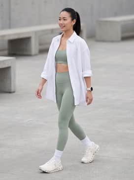 High-waisted compression leggings in a soft sage green paired with a matching sports bra and an oversized white poplin shirt worn open for a chic post-workout look.