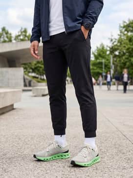 Cloudsurfer 2 – A modern athleisure ensemble consisting of slim-fit black tapered joggers, a crisp white performance polo, and a lightweight lime green windbreaker to echo the shoe's vibrant midsole.