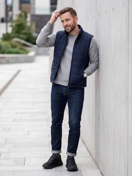 An elevated casual weekend ensemble with dark indigo slim-fit jeans cuffed at the ankle, a high-quality grey marl crewneck sweatshirt, and a navy blue quilted vest.