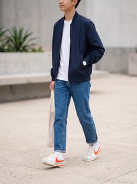 Cortez Textile – Medium-wash straight-leg jeans with a slight cuff at the ankle, a white graphic t-shirt, and a lightweight navy blue bomber jacket for a timeless, casual feel.