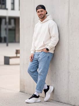 An urban streetwear outfit with relaxed-fit light-wash jeans cuffed at the ankle, an oversized off-white heavy cotton hoodie, and a black snapback cap.