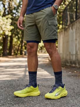 Olive green water-resistant cargo shorts worn with a navy blue long-sleeve base layer and a lightweight beige packable windbreaker for a rugged outdoor aesthetic.