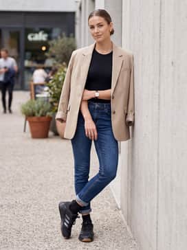 Ankle-length slim-fit jeans rolled once, worn with a black bodysuit and an oversized beige blazer for a high-low fashion mix.