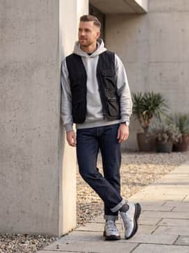 Equipment Agravic Xtr Shoes – A modern streetwear outfit consisting of dark-wash straight-leg denim cuffed at the bottom, an oversized silver-grey hoodie, and a matte black utility vest for a layered, functional aesthetic.