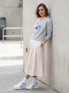 Ex89 – A neutral-toned pleated midi skirt paired with a tucked-in graphic sweatshirt and white crew socks for a trendy 'athleisure-chic' feminine silhouette.