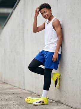 F50 League Mid Cut Firm Ground  Multi Ground Soccer Cleats Kids – Black compression leggings worn under royal blue mesh shorts, paired with a white breathable athletic tank top for high-intensity drill training days.