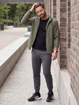 A modern athleisure ensemble consisting of slim-fit charcoal joggers, a fitted black performance tee, and a lightweight olive green windbreaker.