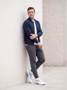 Golf Biom C5 – An urban athleisure outfit pairing charcoal grey tech-fleece joggers with a structured white performance tee and a lightweight navy blue bomber jacket, highlighting the shoe's modern silhouette.
