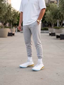 A high-end streetwear outfit consisting of light gray technical joggers, a crisp white oversized heavy-weight tee, and a minimalist gold watch to mirror the shoe's accents.