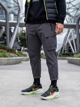 A futuristic streetwear ensemble with charcoal grey multi-pocket cargo pants, a black oversized graphic hoodie, and a technical neon-accented puffer vest.