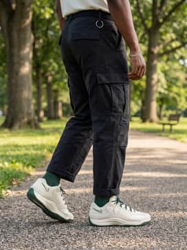 Black cargo pants with multiple utility pockets, a beige windbreaker jacket, and a silver chain necklace for a rugged urban streetwear vibe.