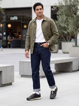 King Indoor Icons Mens Sneakers – Dark indigo straight-leg jeans with a thick cuff at the bottom, a tucked-in white heavyweight cotton tee, and an unbuttoned olive green workwear chore coat.