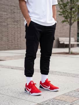 Kobe Ix Elite Low Em Protro – An edgy streetwear ensemble consisting of tapered black cargo joggers, an oversized white graphic t-shirt with red typography, and a black nylon coach jacket.