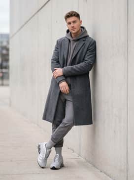 A sleek monochrome outfit consisting of grey tapered joggers, a charcoal grey oversized hoodie, and a long structured wool overcoat for a high-low aesthetic.