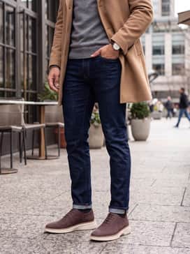 Dark indigo slim-fit jeans worn with a grey merino wool crewneck sweater and a camel-colored overcoat for a refined weekend ensemble.