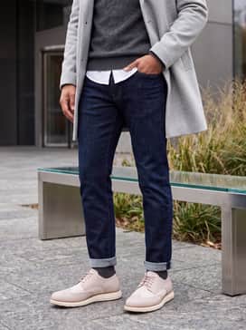 Dark indigo raw denim jeans paired with a charcoal grey merino wool sweater and a light grey overcoat for a polished autumnal look.