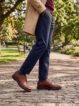 Metropole London – Charcoal grey flannel trousers worn with a burgundy V-neck sweater over a white dress shirt, finished with a classic tan trench coat for a refined scholarly aesthetic.
