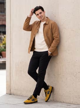 Slim-fit black chinos paired with a cream-colored crewneck sweatshirt and a tan chore coat to create a clean, coordinated outfit that lets the yellow shoes pop.