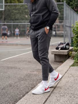 Tapered charcoal grey joggers, a black fitted tech-fleece hoodie, and a red baseball cap that picks up the vibrant hue of the shoe's swoosh.