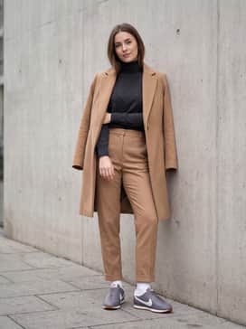 Tailored grey wool trousers and a tucked-in black cashmere turtleneck sweater, topped with a long camel overcoat for a sleek 'clean girl' aesthetic.