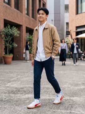 A sophisticated smart-casual look featuring navy blue tailored chinos, a crisp white Oxford button-down shirt, and a tan Harrington jacket that complements the gum sole.