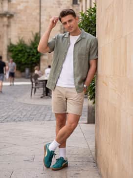 Stone-colored cotton shorts worn with a short-sleeved sage green linen button-up and white crew socks for a trendy, summer-ready look.