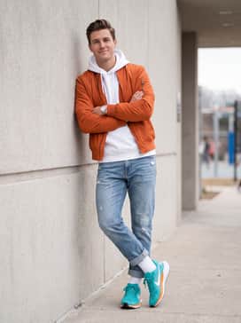 A modern streetwear outfit pairing light-wash distressed denim jeans with a crisp white oversized hoodie and an orange nylon bomber jacket to pull out the accent colors.
