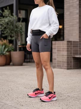 Charcoal grey biker shorts and an oversized white graphic sweatshirt, accessorized with a black technical running belt and a high ponytail for a trendy post-gym coffee run.