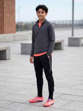 Predator League Firmmultiground Cleats Kids – The High-Intensity Training Look: A charcoal grey moisture-wicking quarter-zip top worn over a vibrant coral base layer, styled with black tapered training pants featuring reflective side detailing.