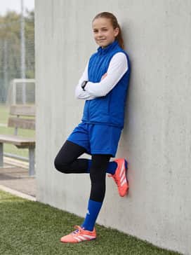 Predator League Firmmultiground Cleats Kids – Cold-Weather Field Gear: A royal blue lightweight insulated vest layered over a white long-sleeve performance tee, paired with black compression tights under navy mesh shorts for a professional athletic look.