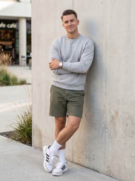 Olive green chino shorts hitting just above the knee, paired with a light grey crewneck sweatshirt and white crew socks pulled mid-calf for a vintage varsity aesthetic that highlights the sneaker's heritage silhouette.