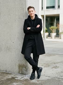 Black tech-fabric joggers tucked into mid-calf athletic socks, a black hooded sweatshirt under a structured trench coat to create a high-fashion, monochrome streetwear silhouette.