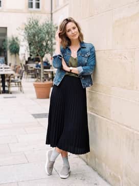 Soft 60 – A black midi-length pleated skirt combined with a tucked-in olive green silk camisole and a denim jacket for a balanced, feminine outfit.