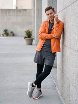 Softride Enzo 5 Wide – Black athletic compression tights layered under loose-fitting charcoal running shorts, topped with a bright orange windbreaker to mirror the shoe's pops of color.