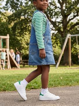 A denim overall dress layered over a green and white striped long-sleeve tee with white ruffle socks for a playful, retro-inspired aesthetic.