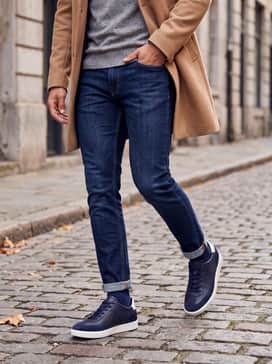 Street Lite – Dark-wash slim-fit denim jeans worn with a grey melange crewneck sweater and a tan overcoat for a polished autumnal weekend ensemble.
