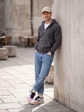 A weekend casual ensemble with tapered light-wash indigo jeans cuffed at the ankle, a navy blue zip-up hoodie, and a beige dad hat that complements the shoe's cream midsole.