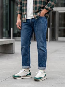 Straight-leg raw denim jeans cuffed at the ankle to showcase the sneakers, worn with a white vintage graphic t-shirt and an unbuttoned plaid flannel shirt in forest green and tan.