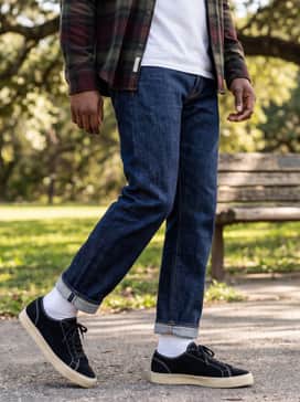 Straight-leg indigo selvedge denim cuffed at the ankle, worn with a white undershirt and an open flannel shirt in earthy tones like burgundy or forest green.