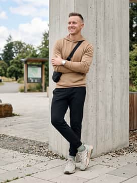 A comfortable travel outfit consisting of black tapered tech joggers, a tan merino wool hoodie, and a sleek black crossbody sling bag.