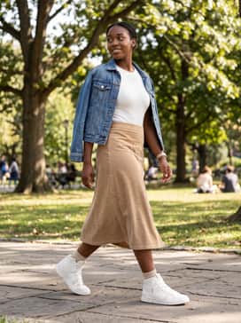 The Roger Clubhouse Mid – A tan corduroy midi skirt, a white rib-knit bodysuit, and a denim jacket draped over the shoulders for a feminine yet sporty ensemble.
