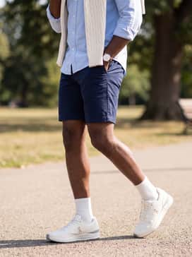 Navy blue chino shorts paired with a light blue oxford button-down and a cream cable-knit sweater draped over the shoulders for a classic preppy vibe.
