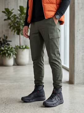 Trailcross Goretex Mountain Bike Shoes – An outdoor adventure ensemble with olive green ripstop cargo pants tucked into the high-top collar, paired with a black thermal base layer and a vibrant orange puffer vest.