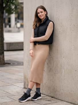 A ribbed knit midi dress in a neutral camel tone layered with a cropped black puffer vest, utilizing the sneakers to ground the feminine silhouette with an athletic edge.