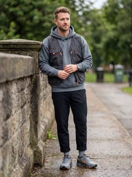 Ua Bandit Trail 3 – A modern gorpcore aesthetic with black tapered tech-wear trousers, an oversized slate grey hoodie, and a utility vest with subtle orange detailing.