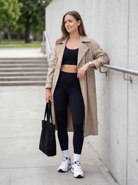 Black high-waisted tech leggings, a matching cropped sports bra, and an oversized beige trench coat for a high-fashion athleisure aesthetic.